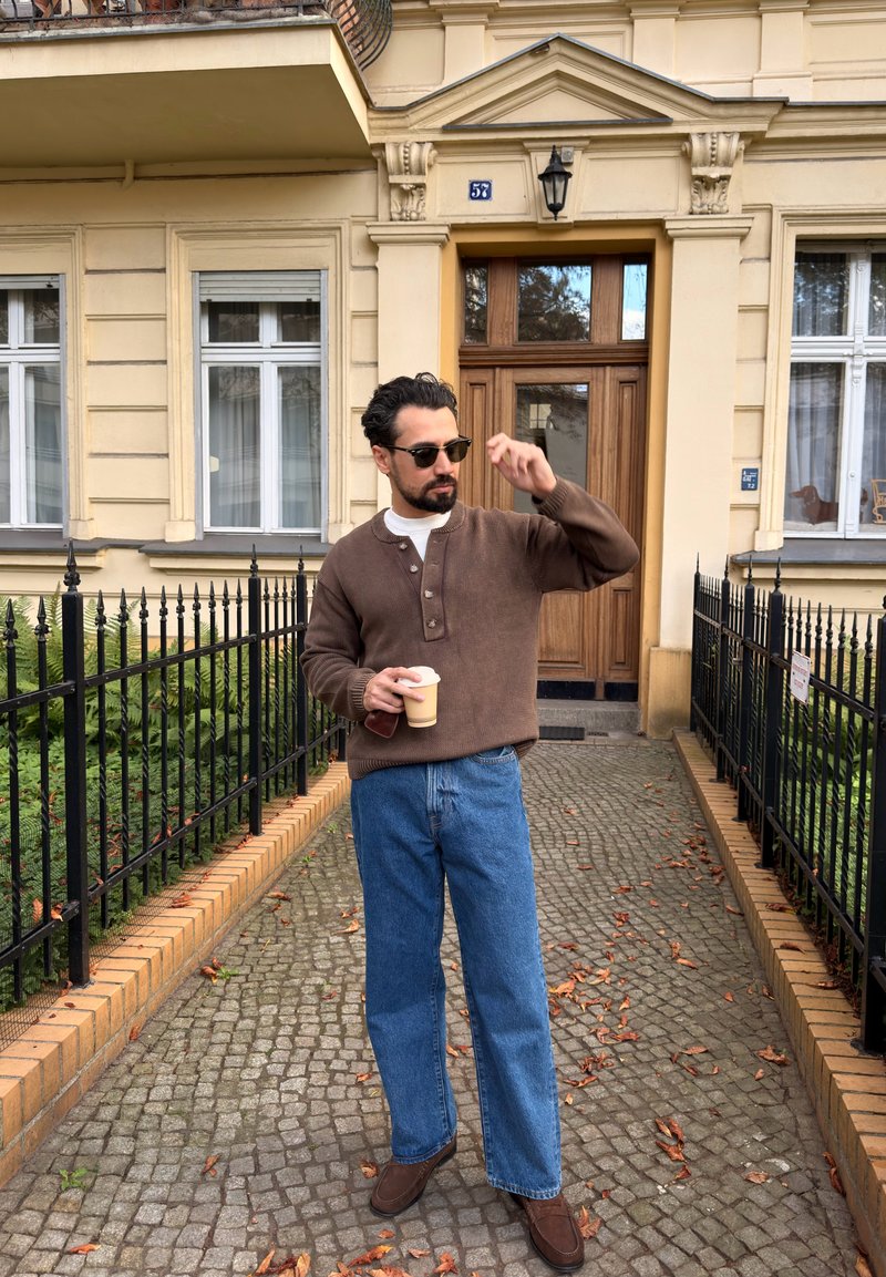 Hombre con gafas de sol y suéter marrón sosteniendo una taza de café, de pie en un camino empedrado frente a la entrada de un edificio beige.