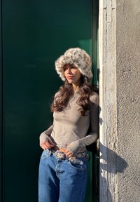 Kunstpelzmütze mit Leopardenmuster, langärmliges beige Oberteil und blaue hoch taillierte Jeans mit gemustertem Gürtel, gelehnt an eine Wand.