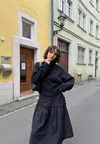 Fekete kötött pulóver magas gallérral, fekete fodros szoknyával párosítva. Fekete táska, az utcán színes épületek háttérben.