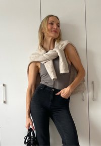 Grå tanktop, lys beige strikkegenser lagt over skuldrene, høy midje svarte jeans, holder en svart veske. Hvit bakgrunn.