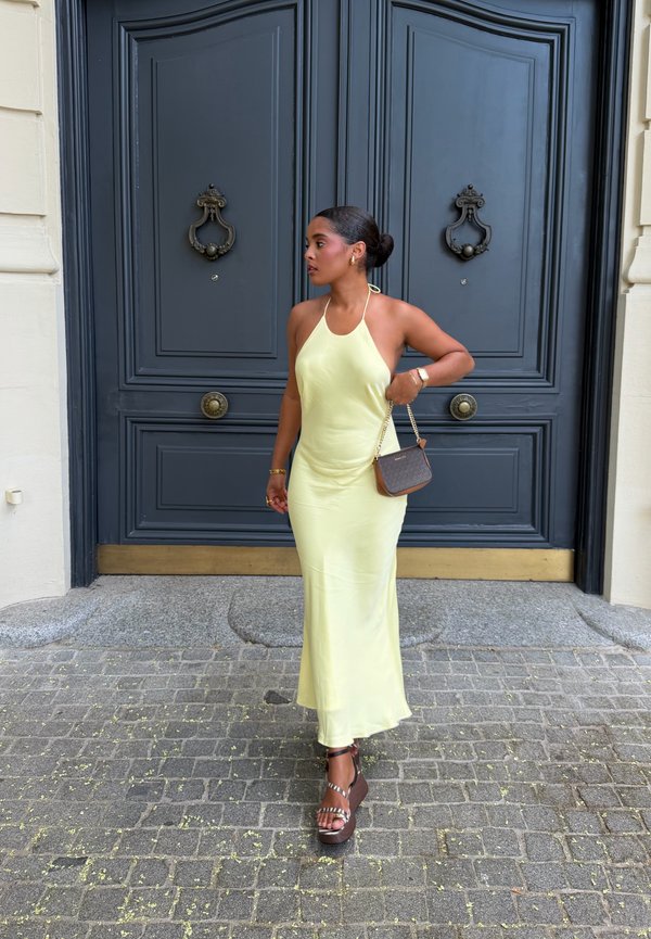 Femme en robe halter jaune pâle et sandales à plateforme se tient sur un pavé en pierre devant de grandes portes doubles bleu foncé avec heurtoirs ornés.