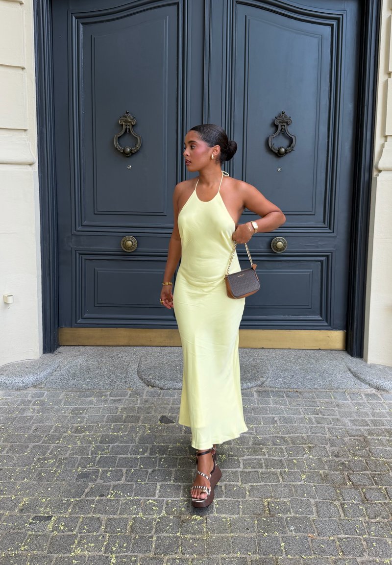 Femme en robe dos nu jaune pâle et sandales à plateforme se tient sur un pavé en pierre devant de grandes portes doubles bleu foncé avec heurtoirs ornés.