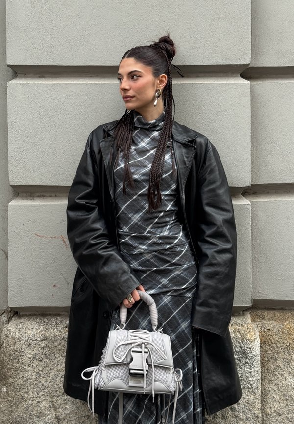 Femme aux cheveux tressés portant un manteau en cuir noir et une robe à carreaux, tenant un sac à main gris, debout devant un mur en pierre.