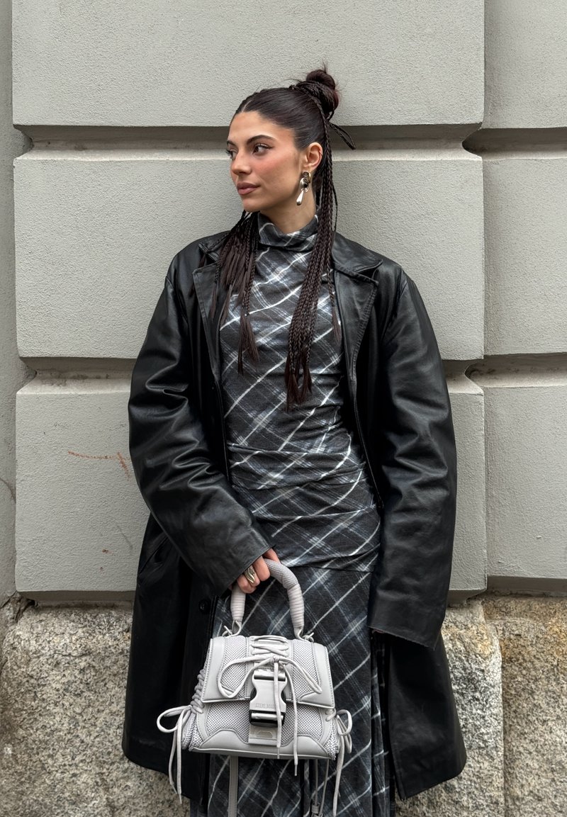 Kobieta z warkoczami, ubrana w czarny skórzany płaszcz i sukienkę w kratę, trzymająca szarą torebkę, stoi na tle kamiennej ściany.