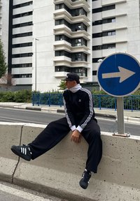 Fekete melegítő együttes fehér csíkokkal, bő szabású, fekete sportcipővel, fehér részletekkel párosítva. Ül egy betonkorláton.