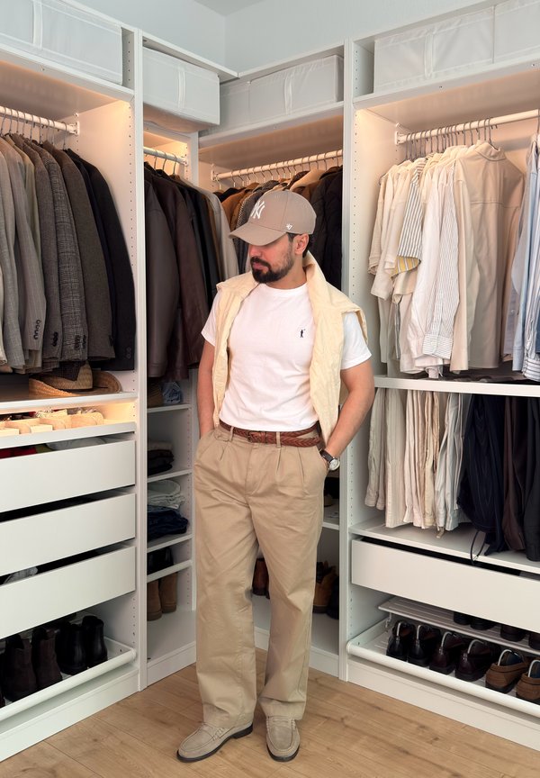 Man wearing beige pants, white t-shirt, and cap stands in a walk-in closet with organized hanging clothes and shelves.