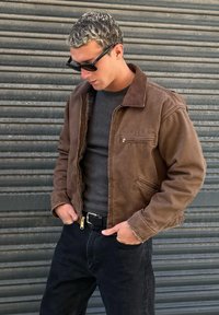 Mann mit brauner Jacke, dunklem Pullover, schwarzer Jeans und Sonnenbrille, stehend mit den Händen in den Taschen vor einem Hintergrund aus Metallrollladen.