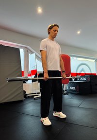 Vállalatot tart mindkét kezében egy fehér pólót és fekete nadrágot viselő személy, aki fekete edzőtermi padlón áll, háttérben edzőgépekkel.