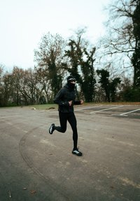 Veste de course noire avec des motifs géométriques, leggings noirs et chaussures noires avec des rayures blanches. Le coureur est sur une surface en gravier, entouré d'arbres.