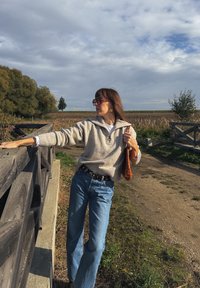 Svetlo siva, teksturirana pulover s ovratnikom, v kombinaciji z modrimi hlačami z visokim pasom in tkano oranžno torbo. Naravna kulisa, oblačno nebo.