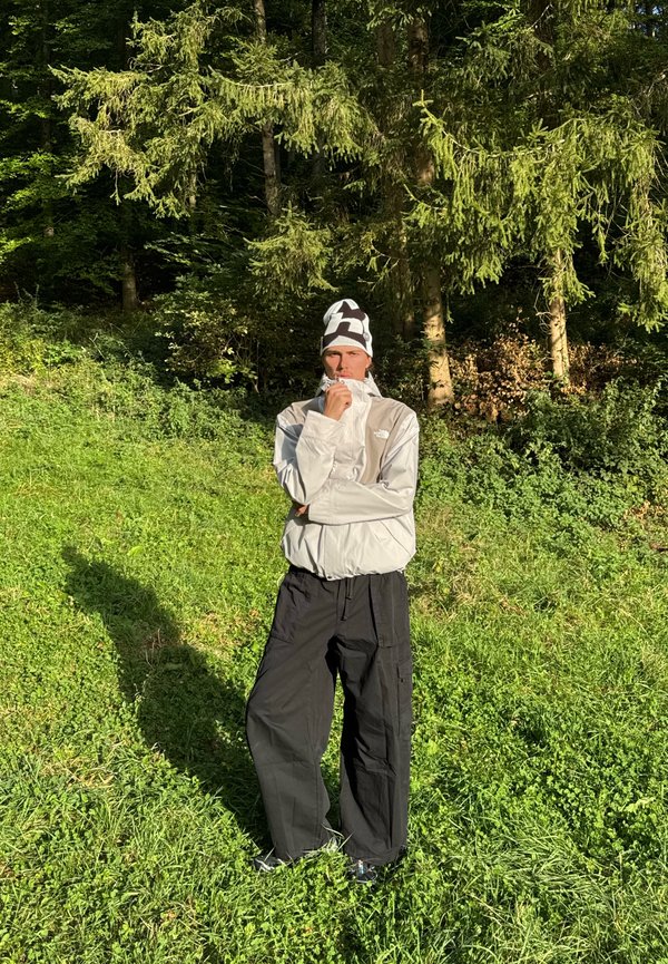Personne portant une veste blanche et un bonnet à motifs, debout sur de l'herbe verte avec des arbres au soleil projetant une ombre au sol.