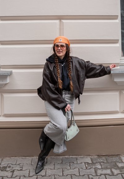 Joven con trenzas largas que lleva una boina naranja, chaqueta de cuero marrón, vaqueros de pierna ancha gris, botas negras y sostiene un bolso verde menta.