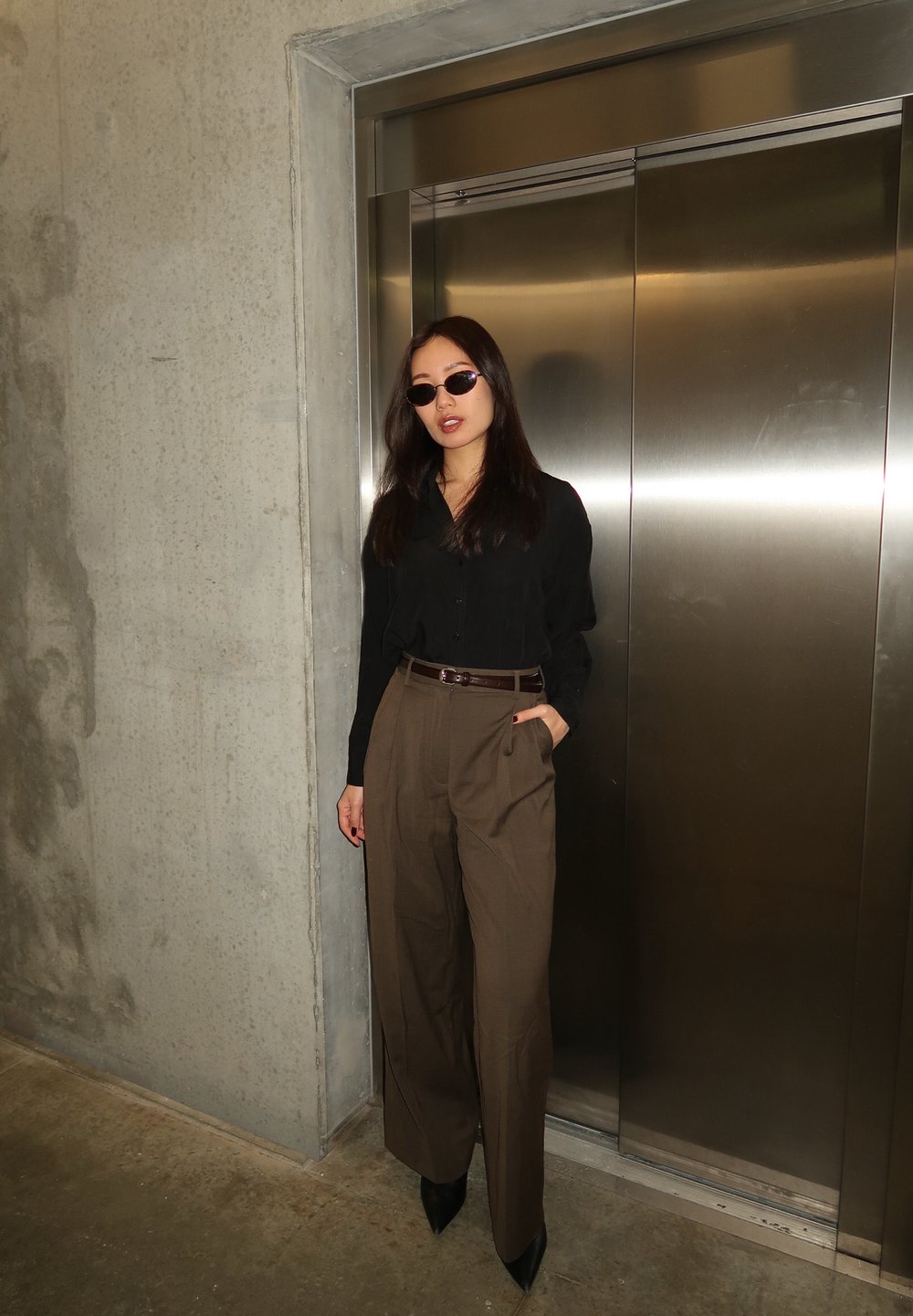 Femme en chemise noire, pantalon plissé marron et lunettes de soleil, appuyée contre un mur à côté des portes fermées d'un ascenseur en acier inoxydable.