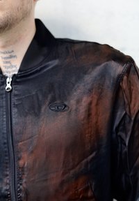 Σκούρο bomber μπουφάν με γκρέιντ ενδιάμεσου καφέ προς μαύρο, διαθέτει φερμουάρ, ριμπ κολάρο και διακριτό λογότυπο στο στήθος.