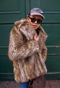 Kabát z kožíšku s kombinací hnědých a šedých tónů, s tlustým límcem a měkkou texturou. Doplňený džínovou čepicí a tmavými slunečními brýlemi.