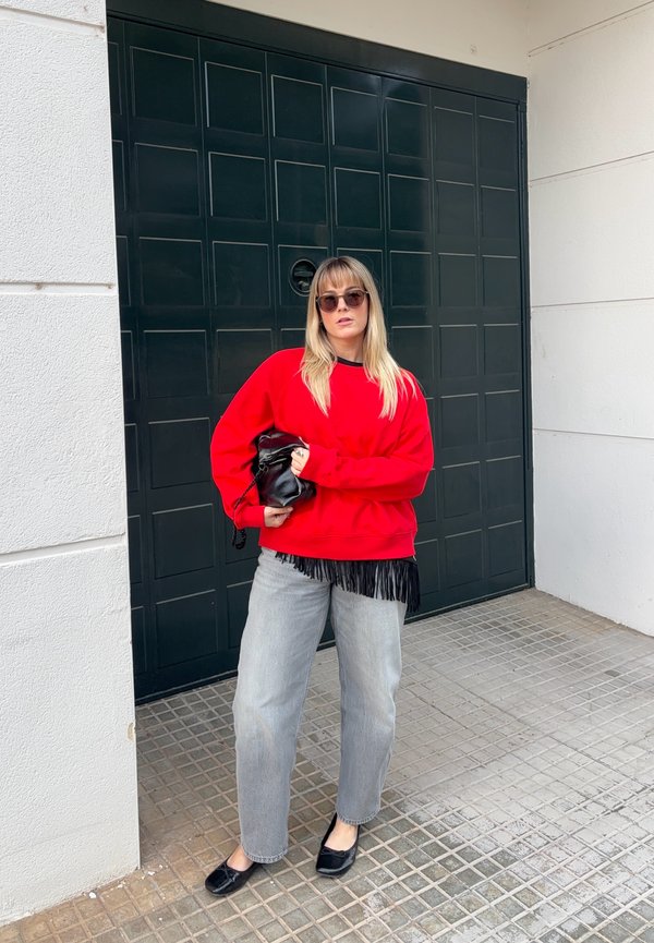 Joven mujer con suéter rojo, vaqueros gris claro, zapatos planos negros y gafas de sol, de pie frente a una puerta de garaje oscura sosteniendo un bolso negro.