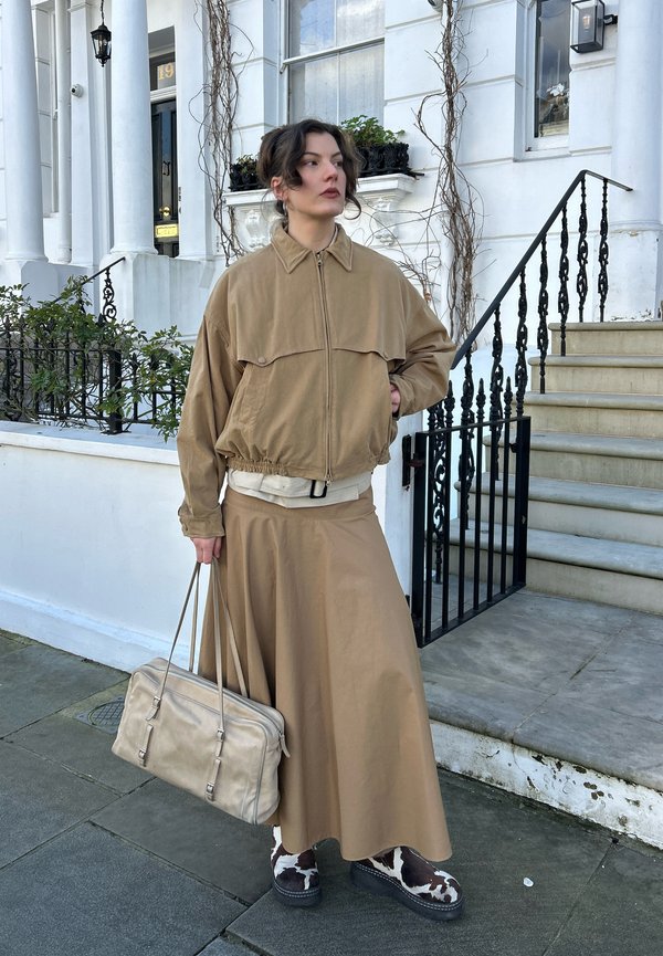 Donna in piedi su un marciapiede con giacca e gonna beige, che tiene una borsa beige, indossando scarpe nere e bianche con motivi, vicino a scale con ringhiera nera.