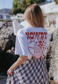 Λευκό μπλουζάκι με κόκκινο κείμενο "HOWDY" και γραφικό σχέδιο με αστέρια και καπέλο καουμπόι. Συνδυασμένο με ένα καρό φούστα. Εξωτερικό περιβάλλον.