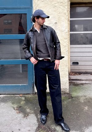 Chaqueta de cuero negra sobre un suéter gris, camisa blanca, pantalones oscuros y zapatos negros. Gorra de béisbol, postura casual, fondo urbano.