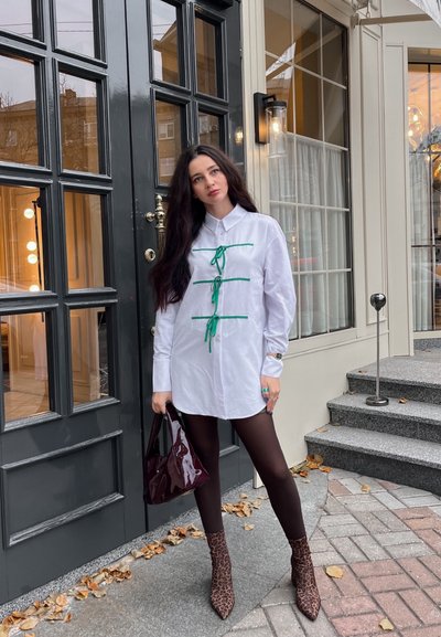 Camisa blanca con detalles verdes, mallas oscuras y botas tobilleras de estampado animal, sosteniendo un bolso burdeos brillante. Entorno al aire libre, escaleras de piedra.