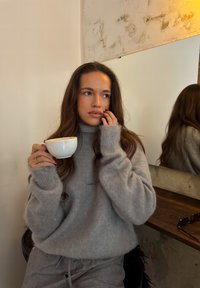 Grå, stor turtlenecktrøje med ribstrikkede manchetter, båret med matchende grå bukser. Hvid keramikkop holdes, der reflekterer minimalistisk indretning.