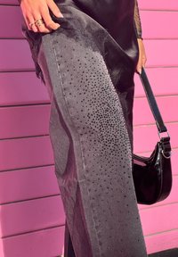 Main tenant une robe en satin noire avec bordure en dentelle, relevée au-dessus d'un pantalon gris orné de petites perles noires, à côté d'un sac à main en cuir verni noir, sur fond de mur rose.