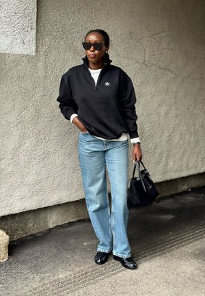 Persona con sudadera negra con cremallera hasta el pecho, vaqueros azul claro, zapatos negros y gafas de sol, sosteniendo un bolso negro, de pie frente a una pared con textura.