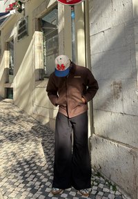 Veste marron à fermeture éclair avec col, pantalon large noir, et casquette beige avec lettrage rouge. Chaussures imprimé léopard, debout sur des pavés.