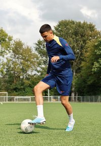Manlig idrottare i en blå långärmad tröja med gula detaljer, blå shorts och färgglada fotbollsskor, som dribblar en vit fotboll på gräset.