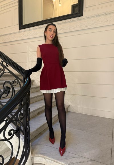 Vestido rojo sin mangas con forma acampanada, fondo blanco en capas; combinado con guantes y medias negras, y zapatos rojos de punta afilada. Escaleras en el fondo.