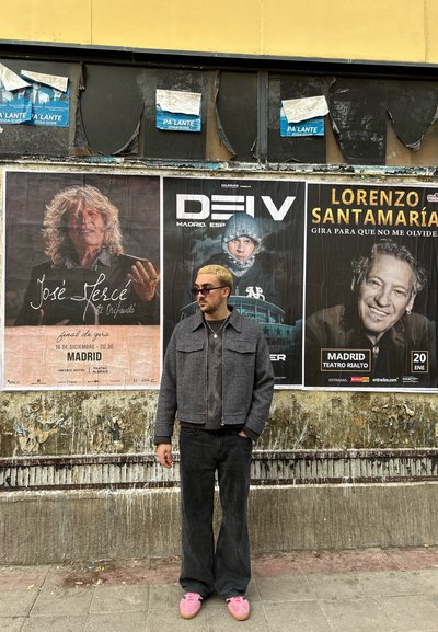 Una persona está de pie frente a carteles desgastados en una pared. Lleva una chaqueta gris, jeans de campana negros y zapatillas rosas.