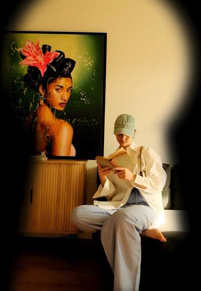 Una persona sentada con una camisa blanca de gran tamaño y pantalones a rayas azul claro lee un libro. Un retrato colorido es visible en el fondo.