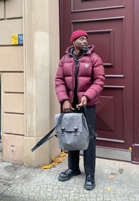 Casaco puffer bordô, camisa preta, calças pretas, mochila cinza mate com alças, sapatos pretos robustos, de pé ao lado de uma porta vermelha com uma placa.