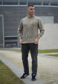 Pull beige en maille torsadée avec poignets côtelés, associé à un jean gris foncé et des baskets noires avec des rayures blanches. Cadre extérieur avec de la verdure.