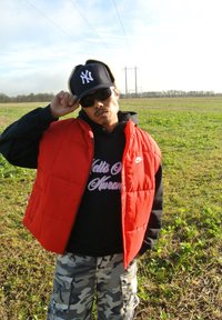 Κόκκινο γιλέκο με φούσκα πάνω από μια μαύρη φούτερ, παραλλαγής cargo παντελόνια, μαύρο καπέλο με λευκό λογότυπο και γυαλιά ηλίου, μπροστά σε φόντο από ανοιχτό πεδίο.