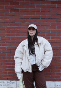 Veste oversize en velours côtelé crème, portée sur un haut en maille blanc. Pantalon marron, casquette beige, et tenant un sac vert clair.