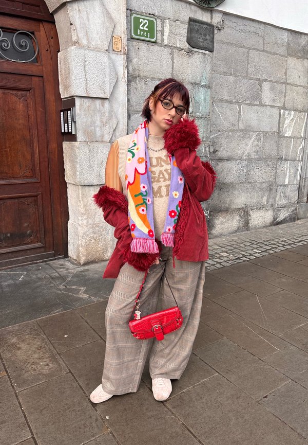 Persona con occhiali posa contro un muro di pietra, indossa una giacca rossa accogliente con bordo in pelliccia, sciarpa decorata, pantaloni larghi a quadri e tiene una borsa rossa.