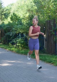 Sportkleding: rode crop top en paarse shorts. Witte sneakers. Hardlopen op een verharde pad, omringd door groen en houten hekken.