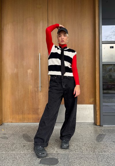 Suéter de punto con rayas en blanco y negro, cuello de tortuga rojo, jeans de pierna ancha negros, calzado negro con agujeros, pendientes de aro plateados, entorno interior.