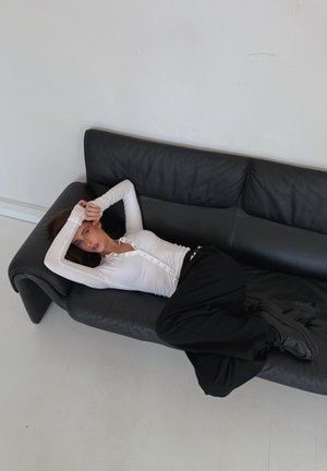 Sofá de cuero negro con una superficie lisa. Persona acostada con una camiseta blanca de manga larga y pantalones negros, manos detrás de la cabeza. Ambiente minimalista.