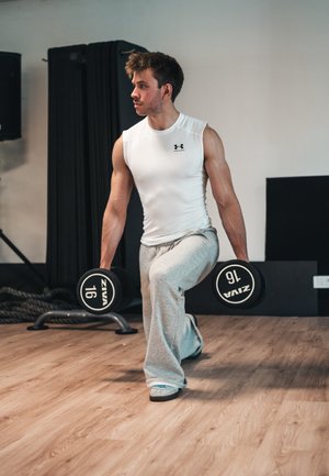 Man in a sleeveless white athletic shirt and grey sweatpants performs a lunge with black dumbbells labeled "16" in a gym setting.