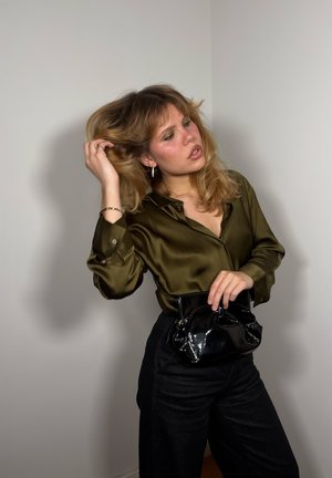 Camisa de satén verde aceituna con cuello puntiagudo, combinada con pantalones negros de talle alto. Sosteniendo un bolso de mano negro brillante.