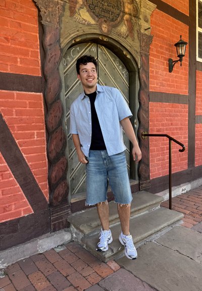 Camisa de cuadros azul claro de manga corta sobre una camiseta negra, pantalones cortos de mezclilla y zapatillas blancas, de pie junto a una puerta de madera ornamental.