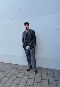 Γκρι διπλόχρωμο σακάκι, ναυτικό γιλέκο, λευκό πουκάμισο και καφέ παπούτσια με λεπτομέρεια κορδόνι. Πλακόστρωτη επιφάνεια με βρύα. Απλός ανοιχτός γκρι τοίχος.