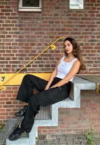 Un débardeur blanc et un pantalon cargo noir sur une personne assise sur un escalier métallique contre un mur en briques, avec une rambarde jaune.