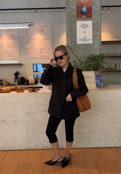 Mujer con blazer negra, pantalones recortados, gafas de sol y bolso marrón de hombro está dentro de una cafetería cerca de un mostrador con pasteles.