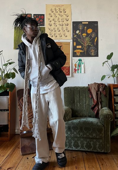 Chaqueta de plumas negra, sudadera gris, pantalones color crema y chanclas negras. Bufanda larga. Sillón verde con un patrón texturizado y suelo de madera.
