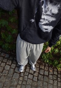 Black hoodie with a graphic design, gray sweatpants, and silver sneakers. Background features green foliage and cobblestone pavement.
