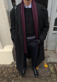 Persona con abrigo largo negro, bufanda morada, guantes negros, pantalones negros y zapatos negros brillantes, de pie sobre una rejilla metálica al aire libre.