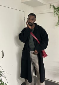 Άνδρας με μαύρο παλτό, γκρι πουκάμισο, ξεθωριασμένο τζιν και κόκκινη τσάντα χιαστί, στέκεται σε εσωτερικό χώρο δίπλα σε λευκή πόρτα και φυτά, αγγίζοντας το πρόσωπό του.